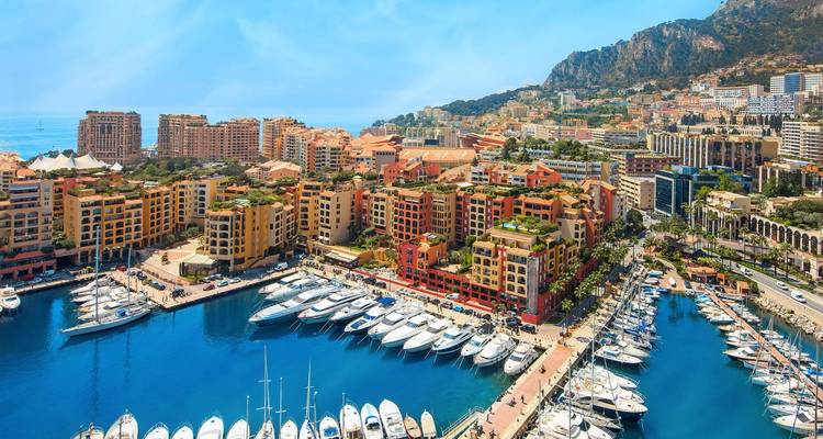 Vue aérienne du Port Hercule de Monaco rempli de yachts de luxe, avec en arrière-plan des bâtiments aux couleurs vives et des montagnes escarpées.