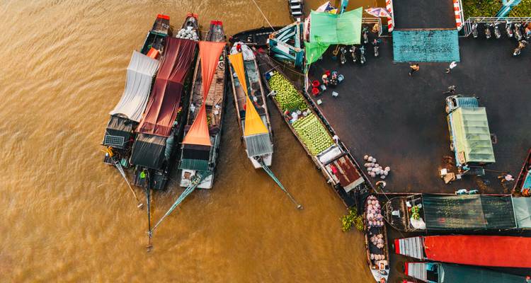Aerial view of vividly canopied market boats moored beside a riverside wharf at dawn.