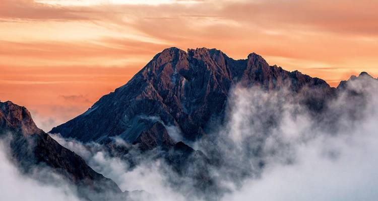 Pic alpin dramatique émergeant au-dessus des nuages tourbillonnants au lever du soleil, teinté de lumière orange.