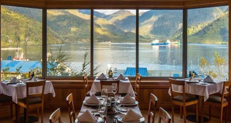 A restaurant dining room with panoramic windows overlooking a calm fjord and mountains.
