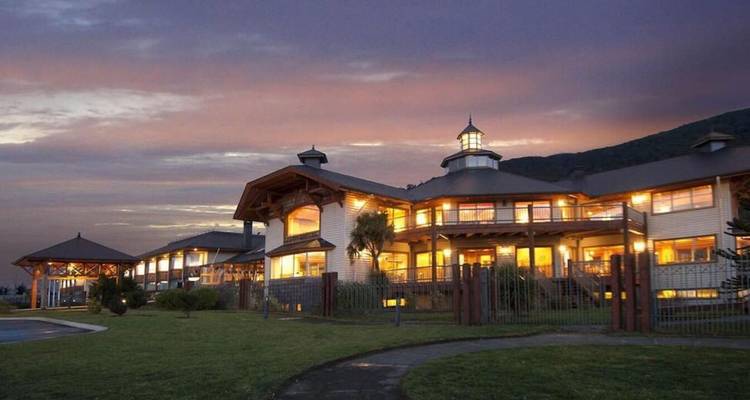 A warmly lit lodge-style hotel against a colorful dusk sky and mountain backdrop.