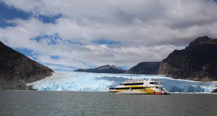 A catamaran approaches a massive blue glacier front in a remote Patagonian fjord.