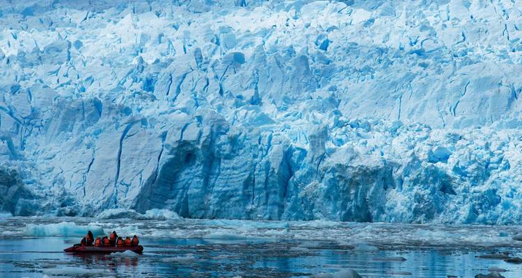 A small inflatable raft with visitors floats beside a towering blue glacier wall.
