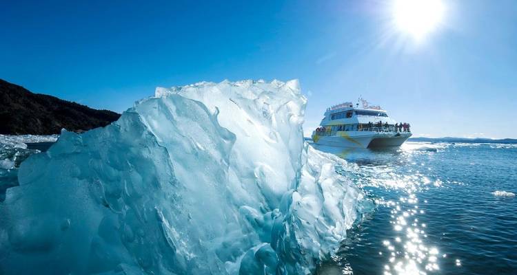 Bright sun glints off the water as an iceberg fragment frames a sightseeing vessel.