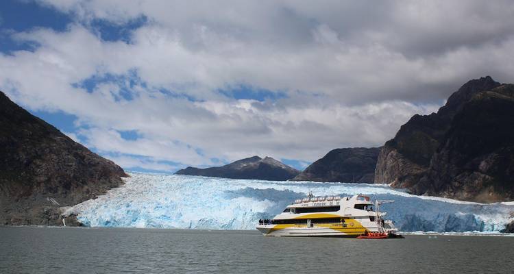 A sightseeing catamaran sails in front of a vast blue glacier with steep mountain walls.