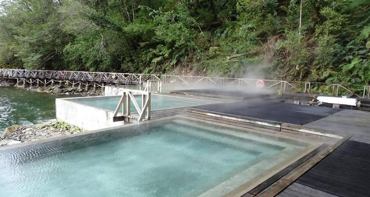 Steaming outdoor thermal pools beside a forested lake with wooden walkways.