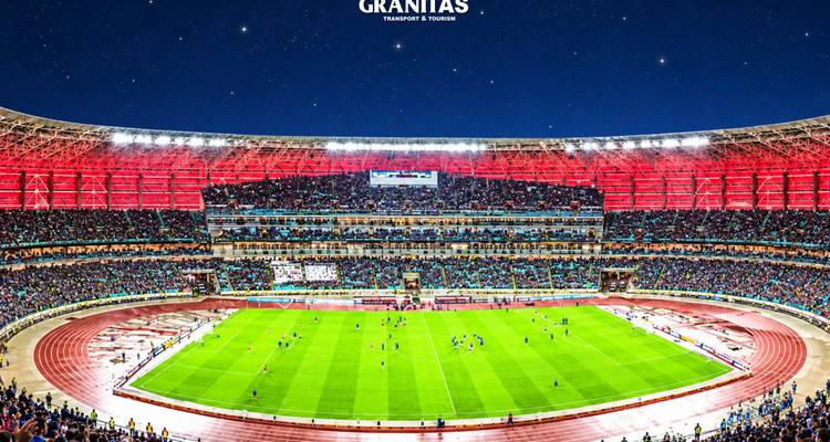 Vue intérieure large d'un stade de football bondé avec un terrain vert et des tribunes rouges sous le ciel nocturne.