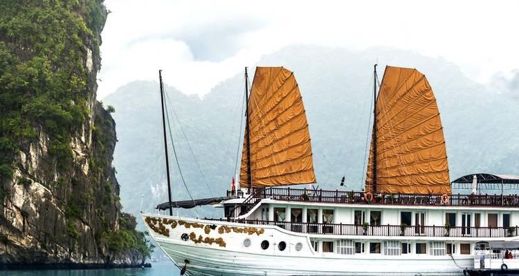 Un barco junco blanco tradicional con velas de color herrumbre se desliza junto a acantilados de piedra caliza escarpada en la Bahía de Halong.