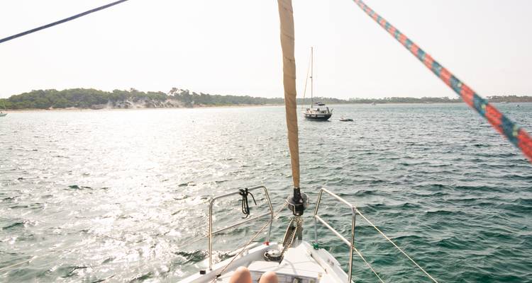 Bow of a sailboat heading toward a sunlit shoreline with another anchored yacht ahead.