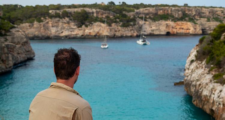 Man looking out from cliffside over a turquoise bay with anchored sailboats.