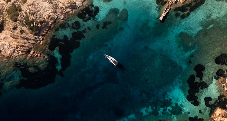 Drone view of a lone sailboat floating in vivid turquoise and navy waters near rugged coastline