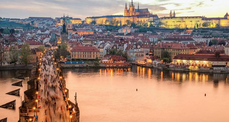 Sunset aerial of Charles Bridge lined with lights, Prague Castle glowing above the city and river.