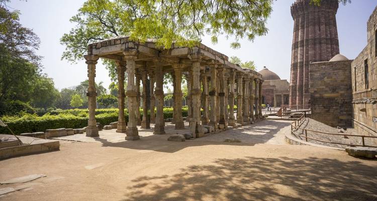 Ancient stone colonnade and courtyard in the Qutub complex shaded by leafy tree.