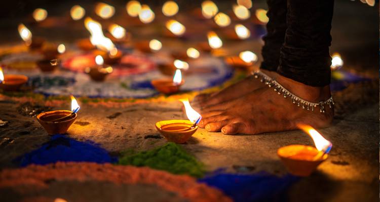 Bare feet with ankle bells stand among glowing oil lamps during a nighttime Diwali ritual.