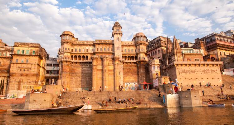 Grand sandstone palace-like ghat with people and boats along the Ganges in warm light.