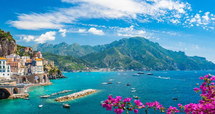 Turquoise Amalfi Coast waters sparkle beneath rugged peaks dotted with boats and magenta flowers.