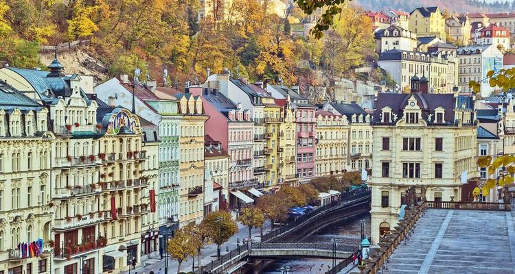 Reihe von pastellfarbenen Spa-Gebäuden und bewaldeter Hanglage mit Blick auf den Kanal in Karlovy Vary während des Herbstes.
