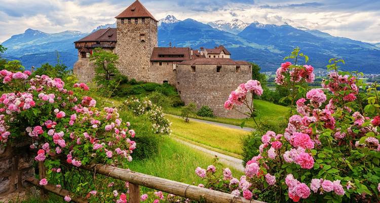 Steinschloss, umrahmt von blühenden rosa Rosen mit Alpengipfeln im Hintergrund.