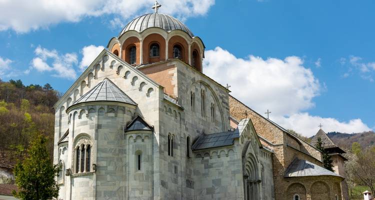 Monastère médiéval serbe aux murs clairs avec nef à dôme situé face à une colline boisée et un ciel bleu.
