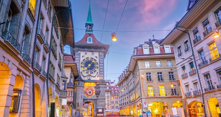 Tour d'horloge historique et rue à arcades illuminées par les lumières chaudes du soir sous un ciel violet.