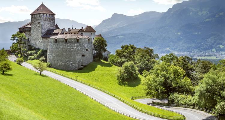 Le château médiéval de Vaduz perché sur une colline herbue avec la vallée alpine et la route sinueuse en contrebas.