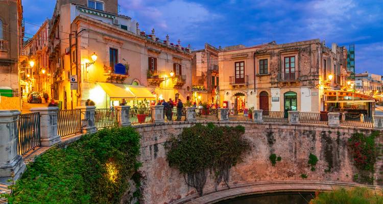 Evening lights illuminate historic stone buildings and bridge over a quiet canal in Syracuse’s Ortigia.