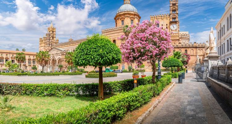 Panoramic garden-lined square and pink flowering trees in front of Palermo Cathedral complex.