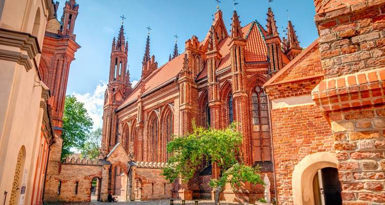 Les flèches gothiques en brique rouge et les arcs-boutants de l'église Sainte-Anne à Vilnius baignés dans la lumière chaude du soleil sur un ciel bleu dégagé.