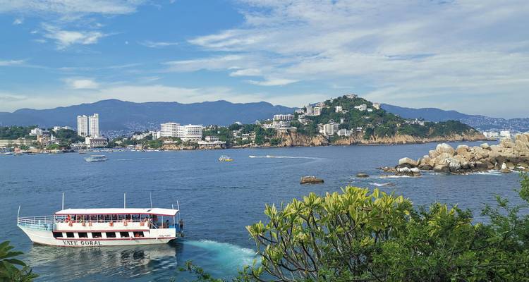 Un bateau de croisière navigue à travers une large baie bleue, avec en arrière-plan la skyline côtière d'Acapulco et des collines verdoyantes luxuriantes.