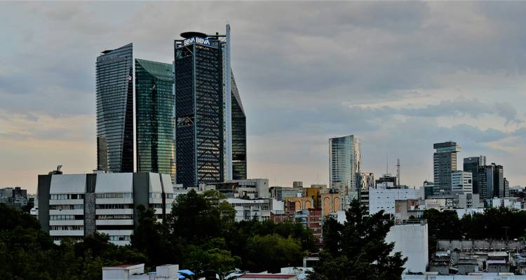 Des gratte-ciel modernes, notamment la tour BBVA revêtue de verre, s'élèvent au-dessus des toits urbains mixtes de Mexico City sous un ciel couvert.