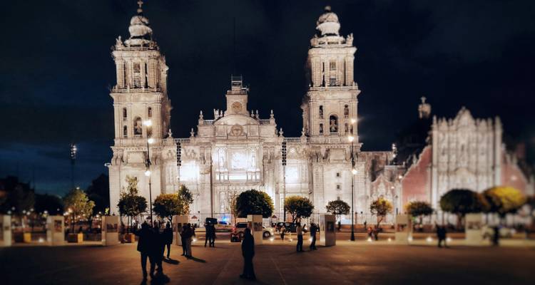 Les tours de la cathédrale de Mexico sont illuminées chaleureusement la nuit, tandis que de petits groupes discutent sur la place principale.