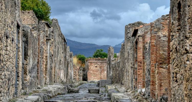 Les rues de pierre usées par le temps de l'ancienne Pompéi mènent vers des montagnes lointaines sous des nuages gris menaçants.
