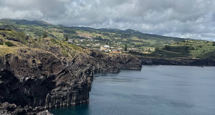 Acantilados volcánicos oscuros que se despeñan en aguas azules tranquilas con tierras de cultivo ondulantes y casas de pueblos en la meseta distante.
