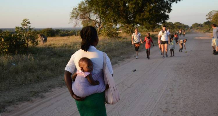 Femme locale avec un bébé sur son dos observe une file de voyageurs marchant le long d'une route rurale poussiéreuse