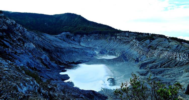 Blue-tinged volcanic crater with steaming white lake surrounded by forested ridges near Bandung.