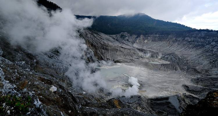 Steaming grey volcanic crater shrouded in mist under a cloudy sky.