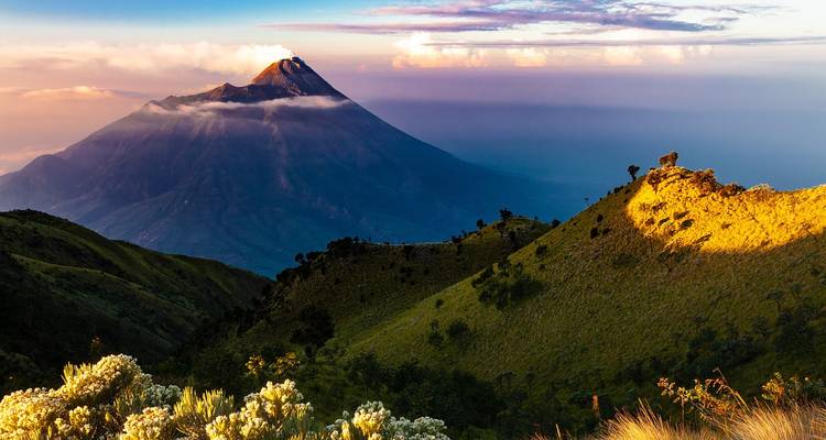 Isolated volcanic cone rising above layers of hills at sunrise with clouds clinging to the slopes.
