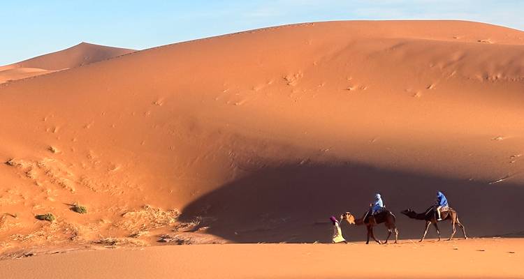 Guide leading a camel with a tourist across sweeping orange dunes casting deep shadows.
