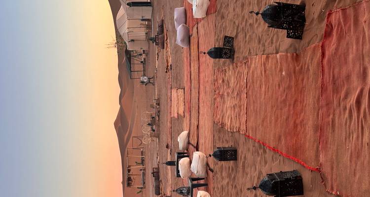Luxury desert camp with rugs, lanterns and tents arranged under a pastel sunset sky.