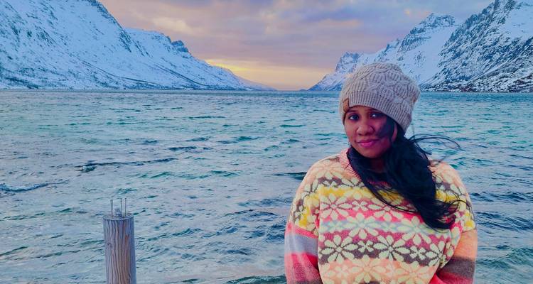 Une femme vêtue d'un pull coloré se tient sur une jetée avec un fjord dramatique et des montagnes s'étendant derrière elle au crépuscule.