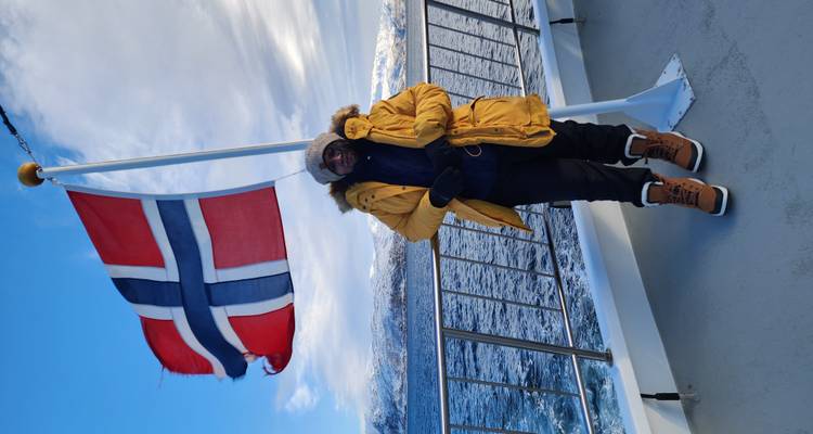 Voyageur en parka jaune se tenant sur le pont d'un bateau à côté d'un drapeau norvégien qui flotte avec une côte enneigée en arrière-plan.