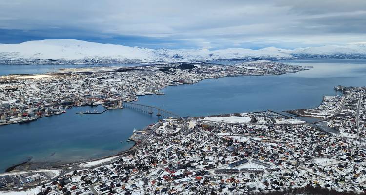 Vue aérienne panoramique de l'île de Tromsø, des fjords environnants et des montagnes enneigées sous un ciel dramatique.