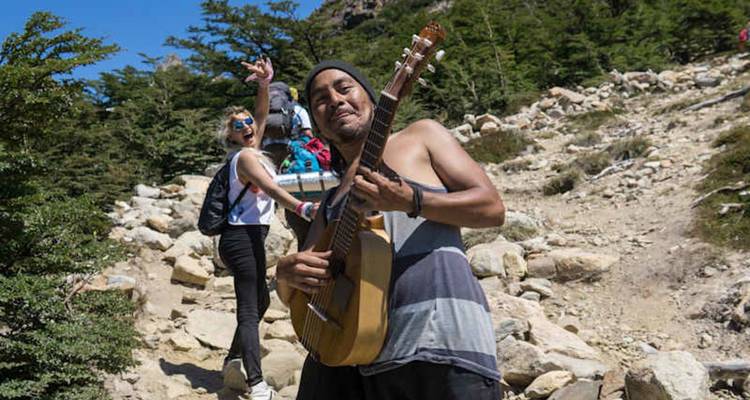 Un músico sin camisa con una guitarra entretiene a los excursionistas en un sendero de montaña rocosa.
