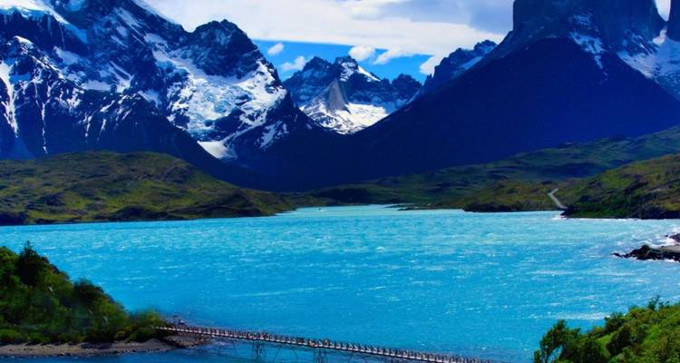 El vibrante lago turquesa Pehoé contrasta con dramáticos picos nevados y un pequeño puente colgante abajo.