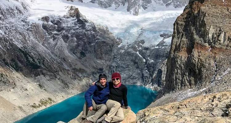 Un couple est assis sur un promontoire rocheux surplombant un lac glaciaire d'un turquoise éclatant, entouré de falaises abruptes et de neige.