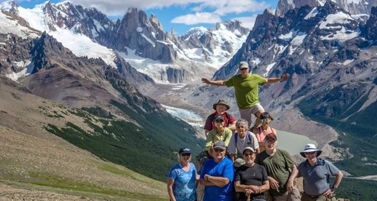 Un grand groupe de randonneurs pose avec enthousiasme sur un versant montagneux avec des pics enneigés spectaculaires et une vallée glaciaire en arrière-plan.
