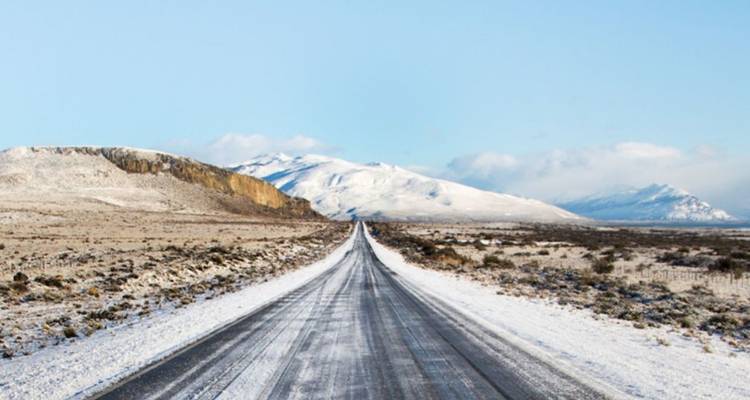 Une longue route droite couverte de neige s'étend vers des montagnes blanches lointaines sous un ciel bleu pâle.