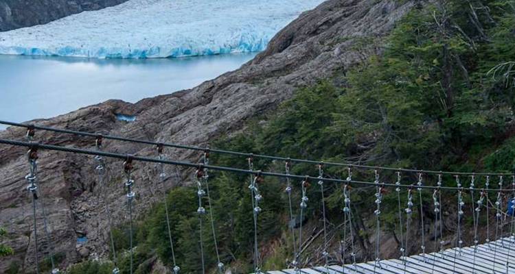 Un pont suspendu enjambe une gorge profonde surplombant un glacier bleu lointain et des versants montagneux escarpés.