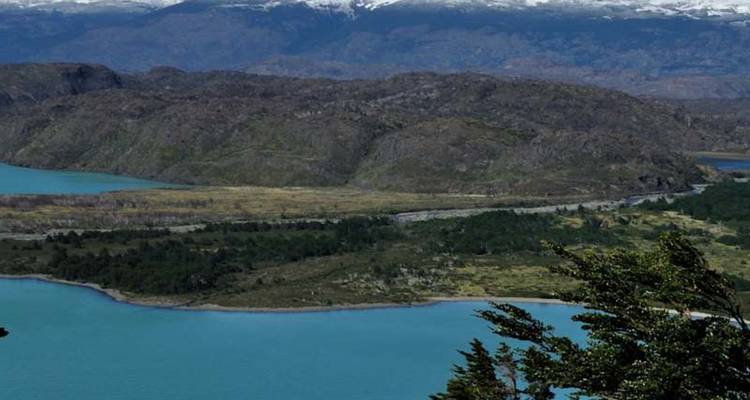 Les eaux turquoise agitées par le vent serpentent entre des collines sombres et des montagnes lointaines aux sommets enneigés sous un ciel nuageux parsemé.