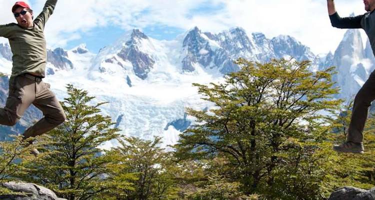 Dos excursionistas llenos de energía saltan en el aire con glaciares de montaña de fondo y arbustos verdes.